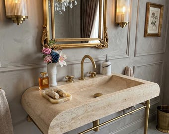 May include: A luxurious bathroom vanity with a beige stone sink and gold fixtures. The sink is set against a gray wall with a gold-framed mirror and sconces. A vase of flowers and perfume bottle add decorative touches.