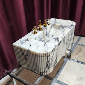 May include: A white marble sink with gold taps, featuring a unique fluted design on the base. The sink is mounted on a metal stand.