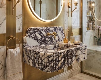 May include: A wall-mounted bathroom sink crafted from white and purple veined marble with a fluted front design. It features a gold-toned metal faucet, a circular backlit mirror above, and two matching gold wall sconces on a brass-accented marble wall.