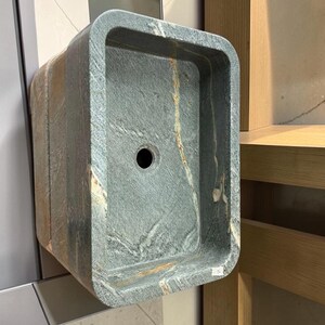 Peut inclure: Un &eacute;vier rectangulaire en mat&eacute;riau semblable &agrave; de la pierre, avec des coins arrondis. L'&eacute;vier est principalement gris avec des stries marron et un trou d'&eacute;vacuation central. Il est mont&eacute; sur un mur, soulignant son design et sa texture uniques.