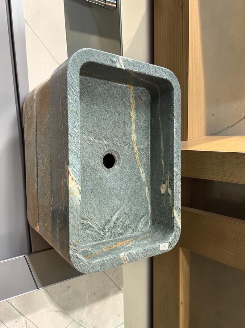 Peut inclure: Un &eacute;vier rectangulaire en mat&eacute;riau semblable &agrave; de la pierre, avec des coins arrondis. L'&eacute;vier est principalement gris avec des stries marron et un trou d'&eacute;vacuation central. Il est mont&eacute; sur un mur, soulignant son design et sa texture uniques.