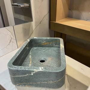 Peut inclure: Un &eacute;vier de salle de bain rectangulaire en pierre aux bords arrondis. L'&eacute;vier est vert mouchet&eacute; avec des stries marron et blanches. L'&eacute;vier a un seul trou d'&eacute;vacuation et est pos&eacute; sur un comptoir blanc.