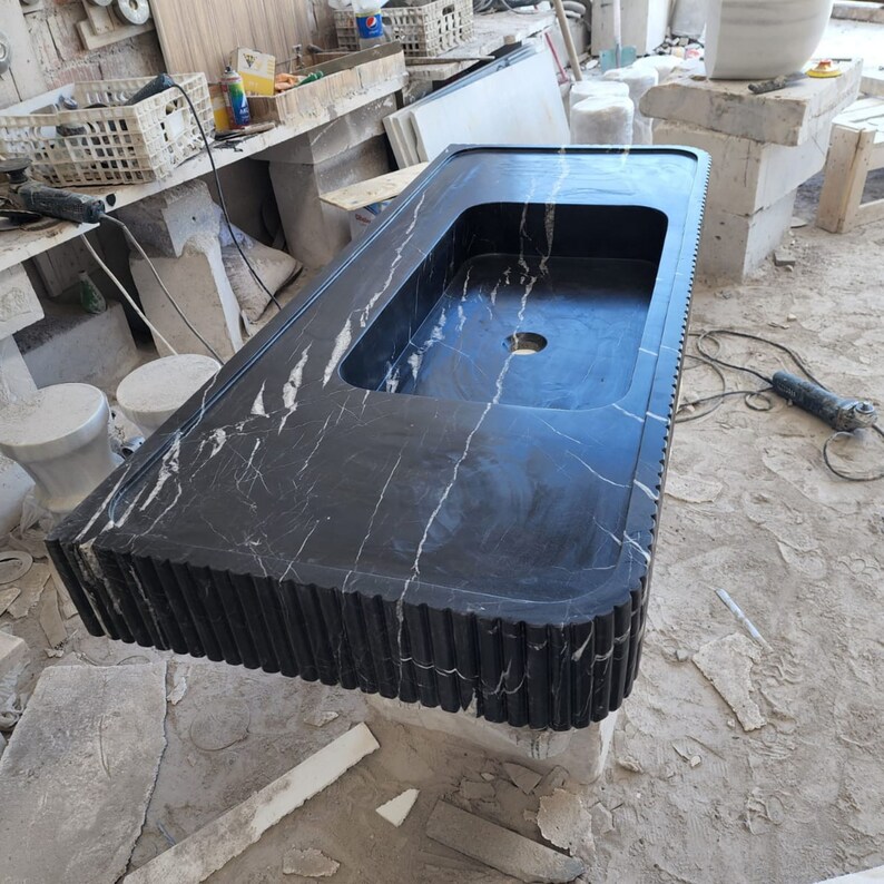 May include: A black marble countertop with a rectangular sink basin and a decorative, fluted edge. The countertop has white veining throughout and a circular drain hole. The piece is in a workshop setting.