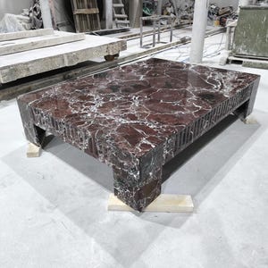 Può includere: Un tavolino rettangolare in marmo rosso scuro con venature bianche. Il tavolo ha una finitura lucida e un design strutturato e scanalato sui lati. Poggia su quattro piccoli supporti in legno, mostrando un'estetica moderna e lussuosa.