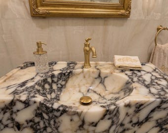 Puede incluir: Un lujoso lavabo de baño con encimera de mármol con grifo dorado, dispensador de jabón y desagüe. Una toalla doblada con bordado dorado descansa sobre la encimera. Un espejo con marco dorado cuelga sobre el lavabo.