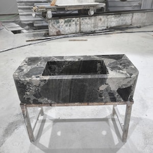 Pode incluir: Uma pia retangular de mármore cinza escuro com uma bacia central, colocada em um suporte de metal prateado. O mármore tem uma aparência natural e padronizada. A pia está em um ambiente de oficina, com máquinas industriais visíveis ao fundo.