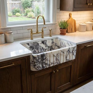 Peut inclure: Un évier de cuisine avec un bassin en marbre et un robinet doré. L'évier est encastré dans un comptoir blanc, et les armoires en dessous sont en bois foncé. Une fenêtre est visible en arrière-plan.