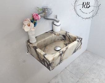 May include: A marble sink with a rectangular basin and a chrome faucet. The sink has a white and gray veined pattern. A small vase with pink and white flowers sits to the left of the faucet. The text "RB Marble Sink" is in a circle on the wall.