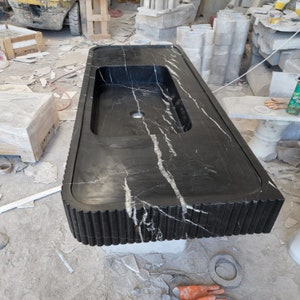 May include: A large, rectangular black marble countertop with a built-in sink basin. The countertop features white veining and a decorative, fluted edge. The sink has a single faucet hole. The countertop is likely for a bathroom or kitchen.