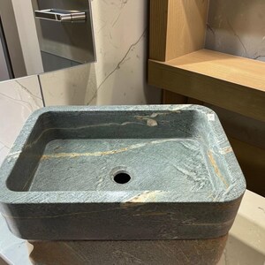 Peut inclure: Un &eacute;vier rectangulaire en pierre, de couleur gris fonc&eacute; et vert. L'&eacute;vier a une forme carr&eacute;e avec des coins arrondis et un trou d'&eacute;vacuation central. La pierre pr&eacute;sente des veines naturelles dans des tons dor&eacute;s et bruns.