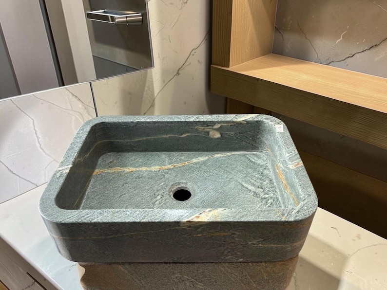 Peut inclure: Un &eacute;vier rectangulaire en pierre, de couleur gris fonc&eacute; et vert. L'&eacute;vier a une forme carr&eacute;e avec des coins arrondis et un trou d'&eacute;vacuation central. La pierre pr&eacute;sente des veines naturelles dans des tons dor&eacute;s et bruns.