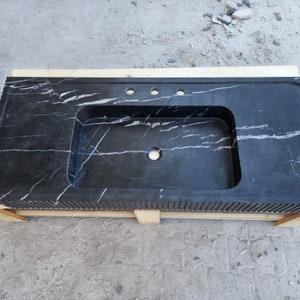 May include: A black marble countertop with a rectangular sink basin and three faucet holes. The countertop has white veining and a decorative edge. It rests on a wooden pallet.