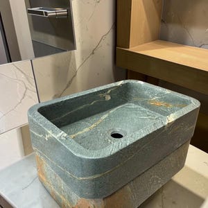 Peut inclure: Un &eacute;vier rectangulaire en pierre, aux tons gris fonc&eacute; et marron. L'&eacute;vier pr&eacute;sente un bassin carr&eacute; et un seul trou d'&eacute;vacuation. Il est fait de pierre en couches et pos&eacute; sur un comptoir blanc.