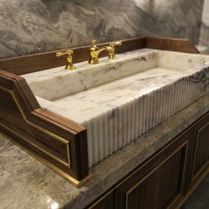 May include: A luxury bathroom vanity featuring a rectangular marble sink and gold taps. The sink is set into a dark wood cabinet with gold trim. The marble has a white and grey veining pattern. The vanity is a statement piece.