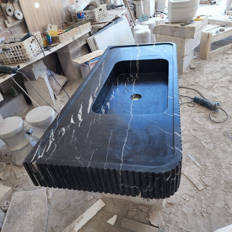 May include: A black marble bathroom sink with white veining. The sink has a rectangular basin with a rounded interior and a single drain hole. The front edge features a vertical grooved design. The sink is in a workshop setting.