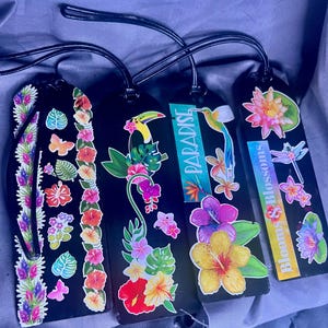 May include: Four black luggage tags with tropical floral and fauna designs. One tag features the word "Paradise" and another reads "Blooms & Blossoms". The tags have black leather straps and are decorated with colorful flowers, butterflies, and a toucan.