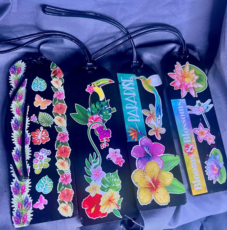 May include: Four black luggage tags with tropical floral and fauna designs. One tag features the word "Paradise" and another reads "Blooms & Blossoms". The tags have black leather straps and are decorated with colorful flowers, butterflies, and a toucan.