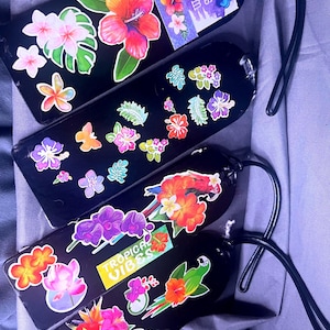 May include: Black luggage tags decorated with colorful floral and tropical sticker designs. One tag has the text "Let Your Dreams Blossom". Another tag has the text "Tropical Vibes". Each tag has a black loop.