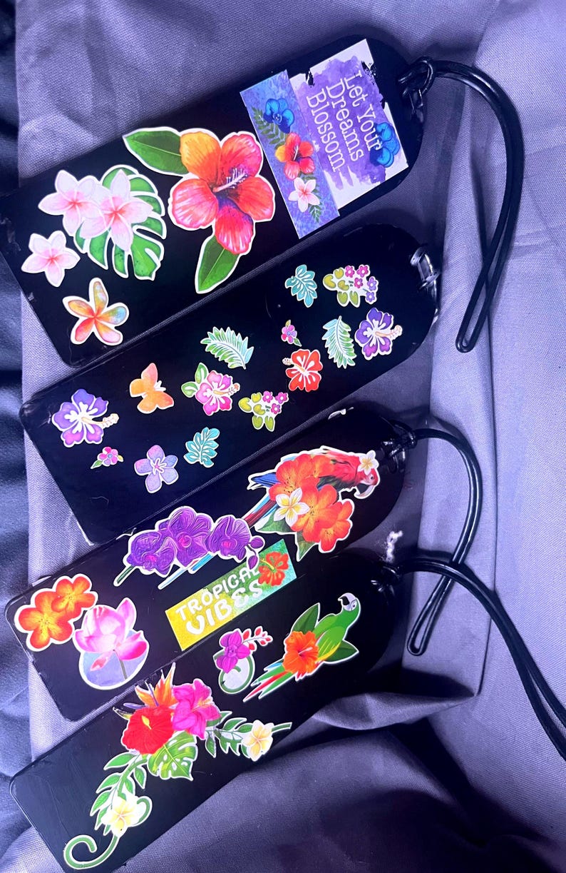 May include: Black luggage tags decorated with colorful floral and tropical sticker designs. One tag has the text "Let Your Dreams Blossom". Another tag has the text "Tropical Vibes". Each tag has a black loop.