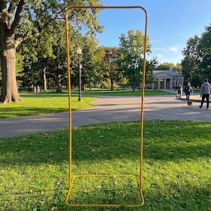 Puede incluir: Un marco metálico amarillo alto de forma rectangular se encuentra sobre la hierba en un parque. El marco tiene una base cuadrada y una parte superior curvada. Hay personas caminando por un sendero pavimentado en el fondo, rodeadas de árboles.