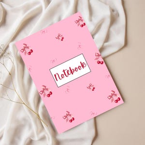 May include: A pink notebook with a cherry and bow pattern. The word "Notebook" is written in red script inside a white rectangle. The notebook is lying on a white fabric.