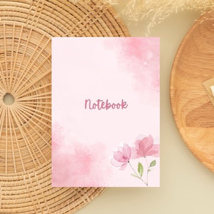 Może przedstawiać: Różowy notatnik ze słowem "Notebook" w kursywie. Okładka ma akwarelowy wzór z różowymi odcieniami i dwiema różowymi ilustracjami kwiatowymi. Notatnik leży na tkanej powierzchni.