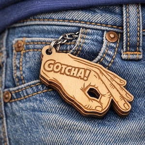 Pode incluir: Um chaveiro de madeira com um design de gesto de mão e a palavra "GOTCHA!". O chaveiro está preso a uma corrente prateada e é colocado em um fundo de ganga azul.