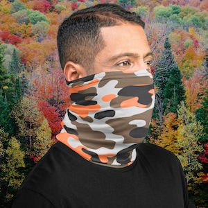 Puede incluir: Una persona lleva una cubierta facial de camuflaje con colores naranja, marrón y blanco. La cubierta facial está hecha de una tela suave y transpirable y está diseñada para ser cómoda de llevar.