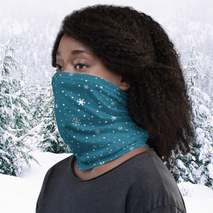 Puede incluir: Una mascarilla facial azul turquesa con un patrón de copos de nieve blancos. La mascarilla la lleva puesta una persona con el pelo oscuro en un entorno invernal nevado.