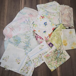 May include: A collection of folded white fabric pieces featuring various floral patterns in shades of pink, blue, yellow, green, and purple. The textiles display diverse botanical prints, including small blossoms, large petals, and leafy designs. Allie's Attic is printed.