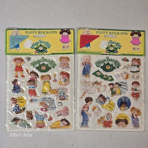 May include: Two sheets of Cabbage Patch Kids Puffy Stick-Ons featuring various cartoon character stickers, text, and illustrations. Each sheet has a yellow header with the brand name and includes multiple colorful adhesive decals of children.