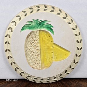 May include: A circular decorative item with a carved pineapple design. The pineapple is split, revealing the textured exterior and the yellow interior with a slice. The piece is framed by a ring with leaf accents, in shades of green and cream.