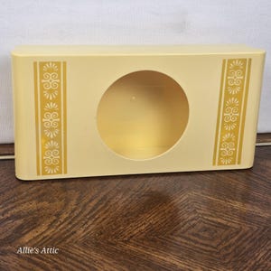 May include: A vintage, rectangular, pale yellow container with a large circular opening. Vertical gold decorative patterns adorn the sides. The container sits on a dark wood surface. The text "Allie's Attic" is visible at the bottom.