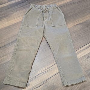 May include: Sage green corduroy trousers with two front pockets and an elasticated waistband. The trousers have a relaxed fit and are made of a soft, ribbed fabric. Suitable for children.