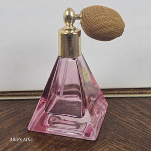 Pode incluir: Um frasco de perfume de cristal rosa com um atomizador dourado e uma pêra bege. O frasco em forma de pirâmide tem um gargalo dourado. O rótulo no frasco diz "SC Crystal". O frasco está sobre uma superfície de madeira.