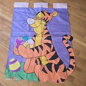 May include: A purple fabric wall hanging featuring a cartoon tiger holding Easter eggs. The tiger is orange with black stripes and yellow accents. The eggs are decorated with various patterns and colors. The top has pink tabs for hanging.