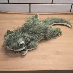 May include: A plush, green stuffed alligator toy with a shaggy texture. The toy has a white belly and a long tail. The alligator is lying on a wooden surface.