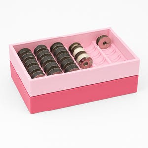 Peut inclure: Une boîte à couture à deux niveaux, rose et rouge, remplie de bobines de fil. Le plateau supérieur, rose clair, contient plusieurs bobines de fil gris foncé et quelques bobines blanches et argentées. Le plateau inférieur est rouge plus foncé.