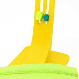 May include: A bright yellow display stand holds two small, colorful earrings: one lime green and one teal. A neon green zipper is laid across the base of the stand. The background is white.