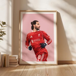 May include: Framed art print featuring a stylized illustration of a soccer player in a red jersey with white stripes and the number 8. The jersey has the "Standard Chartered" logo. The background is a soft pink.