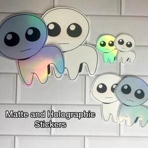 May include: A set of four holographic and matte stickers featuring a cartoon character with large eyes and a simple design. The stickers are in various sizes and colors, including blue, green, and white.