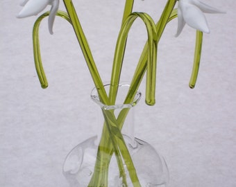 May include: A bouquet of three white glass snowdrop flowers with green stems in a clear glass vase with a round, textured body.