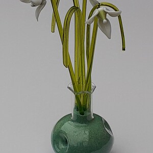 Può includere: Un vaso di vetro con una superficie verde testurizzata e un collo di vetro trasparente contiene un mazzo di fiori di bucaneve di vetro bianco con steli di vetro verde.