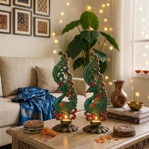 May include: Two peacock candle holders with green, red, and gold detailing. The candle holders are on a wooden table with other decorative items. The background includes a sofa, plant, and fairy lights.