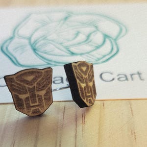 Puede incluir: Un par de pendientes de madera con el símbolo Autobot. Los pendientes son marrones con un diseño marrón claro. El fondo está borroso, con la palabra "Cart" visible.