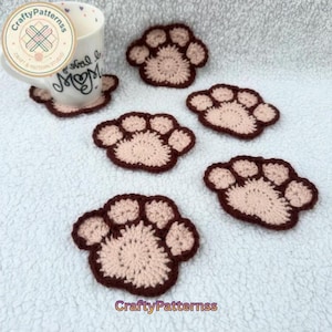 Peut inclure: Ensemble de six dessous de verre faits à la main en crochet en forme de pattes de chien, marron et beige clair. Une tasse blanche avec le mot "Mom" et un dessous de verre assorti sont également visibles. Le logo "CraftyPatternss" est dans l'image.