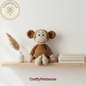 Könnte beinhalten: Ein handgehäkelter Affe in Braun- und Cremetönen sitzt auf einem Holzregal. Der Affe hat große Ohren, ein lächelndes Gesicht und cremefarbene Hände und Füße. Das CraftyPatternss-Logo ist sichtbar.