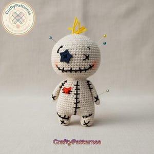 May include: A handmade crochet doll with a cream-colored body, a stitched black mouth, and a blue star-shaped eye patch. The doll has a red heart stitched on its chest and yellow hair. Several pins are inserted into the doll's head and body. The text "CraftyPatternss" is at the bottom.