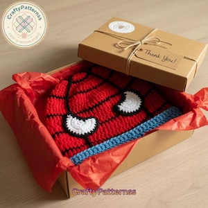 May include: A red, black, white, and blue crocheted Spider-Man hat inside a cardboard box lined with red tissue paper. The box is tied with twine and has a tag that says "Thank You!". The hat is a handmade item.