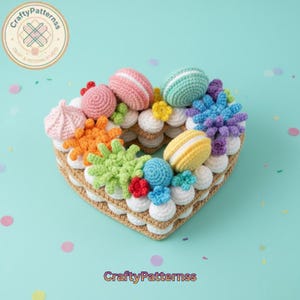 Op de afbeelding: Een hartvormige gehaakte taart met kleurrijke garendecoraties. De taart heeft roze, groene, gele en blauwe macarons, samen met andere gehaakte versieringen. De basis is lichtbruin en de tekst "CraftyPatternss" is zichtbaar.